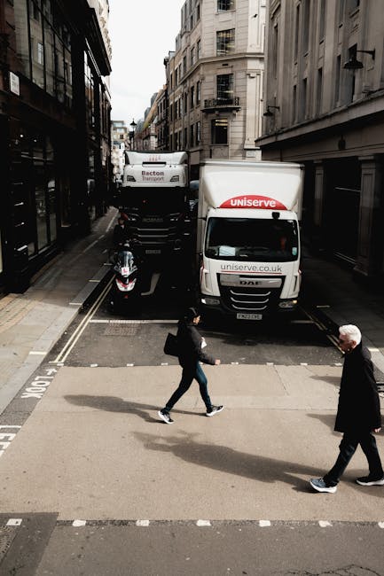A street scene in an urban area with narrow roads lined by multi-storey buildings featuring classic architectural details. In the foreground, two pedestrians are crossing the road; a woman dressed in dark clothing with a backpack and a man in a dark coat and white hair. Behind them, a white van with the 'Uniserve' logo and website address is parked partly on the pavement near a delivery truck marked 'Becton Transport.' The van is positioned close to the building entrances, with the loading area visible where furniture and boxes might be staged for home relocation purposes. A scooter is parked near the sidewalk, and the street is clean with markings for pedestrians. The scene captures a typical city environment involved in moving activities, with the presence of logistics vehicles supporting furniture transport and packing processes related to house removals, as highlighted on the webpage about Kingston removals quotes.
