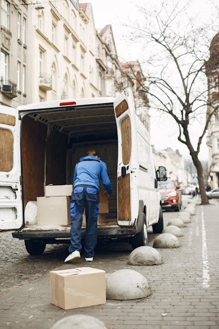 A street scene in an urban area with narrow roads lined by multi-storey buildings featuring classic architectural details. In the foreground, two pedestrians are crossing the road; a woman dressed in dark clothing with a backpack and a man in a dark coat and white hair. Behind them, a white van with the 'Uniserve' logo and website address is parked partly on the pavement near a delivery truck marked 'Becton Transport.' The van is positioned close to the building entrances, with the loading area visible where furniture and boxes might be staged for home relocation purposes. A scooter is parked near the sidewalk, and the street is clean with markings for pedestrians. The scene captures a typical city environment involved in moving activities, with the presence of logistics vehicles supporting furniture transport and packing processes related to house removals, as highlighted on the webpage about Kingston removals quotes.