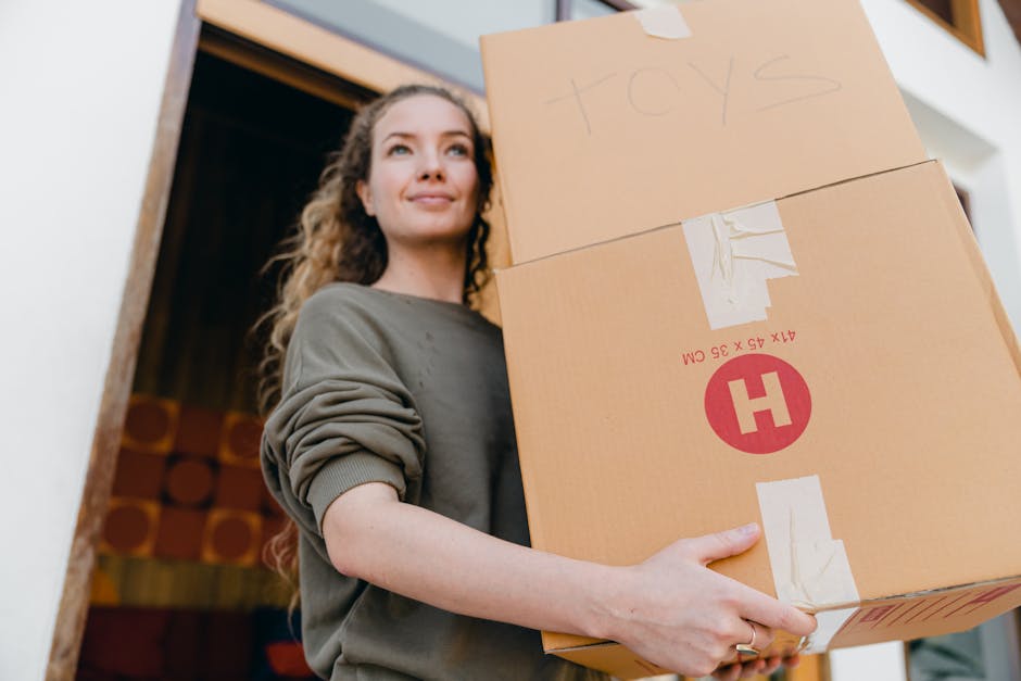 A woman with long, curly hair and a neutral expression stands inside a home near a doorway, holding three cardboard boxes stacked one on top of another. The top box is labeled 'toys' with handwritten text, while the other boxes feature printed labels and symbols, including a red circle with a white 'H' and measurements. She is wearing a grey sweatshirt with the sleeves rolled up, and she is grasping the boxes securely with both hands. The background shows part of the interior, including a wooden doorframe and a wall with a decorative pattern made of stacked wooden logs. This image depicts the process of packing or moving, with the woman engaged in home relocation activities, supported by a professional removals service such as [COMPANY_NAME], preparing for furniture transport and home removal logistics.
