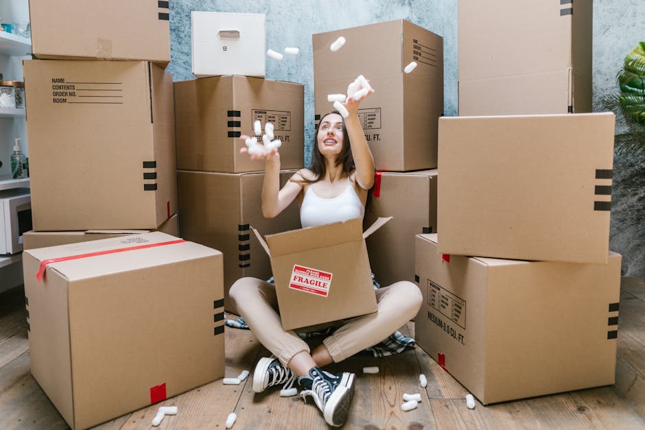 A young woman with long dark hair, sitting cross-legged on a wooden floor indoors during a house removal process, surrounded by various large cardboard boxes, some labeled 'fragile' and sealed with red tape. She is wearing a white tank top, beige pants, and black sneakers with white soles, and appears to be enjoying herself as she throws packing foam peanuts into the air. The boxes are stacked against a textured light blue wall, and there is a partial view of a kitchen counter with a microwave on the left side. Several foam peanuts are scattered around her on the floor, and she is engaging in packing or unpacking activities as part of a home relocation handled by [COMPANY_NAME], focusing on furniture transport and loading procedures during the moving day, aligned with the Kingston area moving tips outlined on the page.