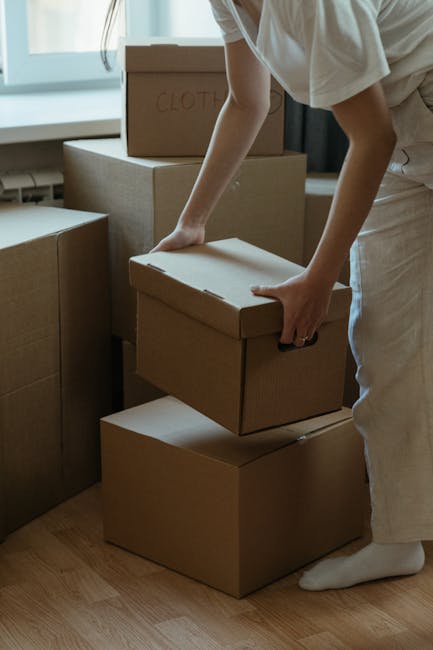 A woman with long, curly hair and a neutral expression stands inside a home near a doorway, holding three cardboard boxes stacked one on top of another. The top box is labeled 'toys' with handwritten text, while the other boxes feature printed labels and symbols, including a red circle with a white 'H' and measurements. She is wearing a grey sweatshirt with the sleeves rolled up, and she is grasping the boxes securely with both hands. The background shows part of the interior, including a wooden doorframe and a wall with a decorative pattern made of stacked wooden logs. This image depicts the process of packing or moving, with the woman engaged in home relocation activities, supported by a professional removals service such as [COMPANY_NAME], preparing for furniture transport and home removal logistics.