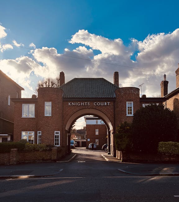 A wide shot of a residential street featuring the entrance to Knights Court, a brick building with an arched gateway in the centre, branded with the name 'Knights Court'. The gateway leads to a driveway with parked cars visible in the distance. On either side of the entrance, there are two small, landscaped areas with bushes. The building has a tiled, dark roof, and multiple windows, some with white frames. In front of the building, the street is paved with asphalt, with curbs and a pavement on both sides. The sky above is partly cloudy with patches of blue, and sunlight casts shadows on the building and street. The scene depicts a quiet area suitable for house removals and furniture transport during a home relocation process, managed by [COMPANY_NAME], with the focus on efficient loading and moving logistics.