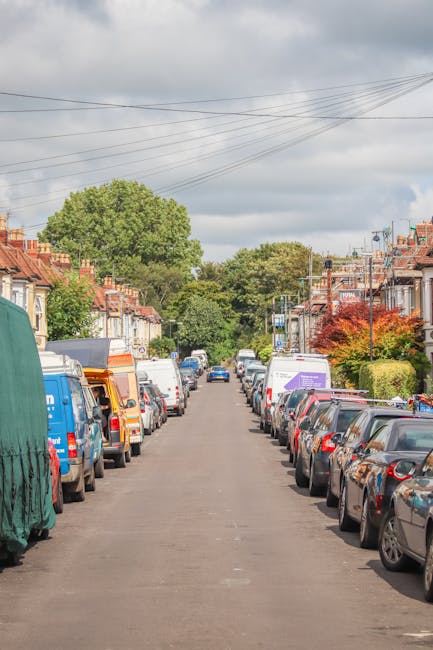 The image shows a residential street during daytime with a row of parked cars lining both sides of the pavement. In the foreground, a large vehicle from [COMPANY_NAME], likely a removal van, is partially visible on the left side, covered with a green protective tarp. The street appears to be a typical urban area with terraced houses, some with small front gardens, and a mix of trees and shrubbery adding greenery to the scene. Overhead, wires stretch across the sky, which is partly cloudy with patches of sunlight illuminating the street. The scene likely captures a moment during a home relocation or moving process, with parking permits visible for the area, relevant to the Kingston upon Thames location specified in the page title. The image illustrates the logistical environment for furniture transport and packing activities associated with house removals along Clarence Street and Eden Street, highlighting parking arrangements and vehicle positioning essential for efficient moving operations by [COMPANY_NAME].