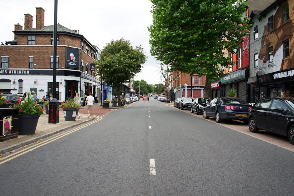 A wide shot of a residential street featuring the entrance to Knights Court, a brick building with an arched gateway in the centre, branded with the name 'Knights Court'. The gateway leads to a driveway with parked cars visible in the distance. On either side of the entrance, there are two small, landscaped areas with bushes. The building has a tiled, dark roof, and multiple windows, some with white frames. In front of the building, the street is paved with asphalt, with curbs and a pavement on both sides. The sky above is partly cloudy with patches of blue, and sunlight casts shadows on the building and street. The scene depicts a quiet area suitable for house removals and furniture transport during a home relocation process, managed by [COMPANY_NAME], with the focus on efficient loading and moving logistics.