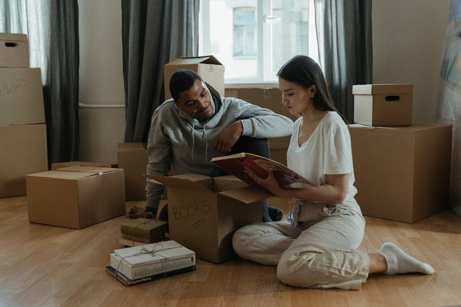 A young woman and a man are inside a room with light-colored wooden flooring, preparing for home relocation. The woman is sitting on the floor, wearing light-colored casual clothing and white socks, holding an open book. The man is kneeling beside her, dressed in a grey hoodie, observing the book. Surrounding them are several cardboard boxes, some sealed and labeled, with handwritten notes such as 'BOOKS,' and additional packaging materials like wrapped books tied with string. The boxes vary in size and are arranged around them, some stacked against the wall and others on the floor, indicating the packing process. The room has a window with natural light illuminating the scene, with dark curtains pushed aside. The environment suggests an active moving or packing process, which [COMPANY_NAME], such as a professional removals service, may facilitate during house relocations in Kingston, Kingston upon Thames, involving careful packing and furniture transport.