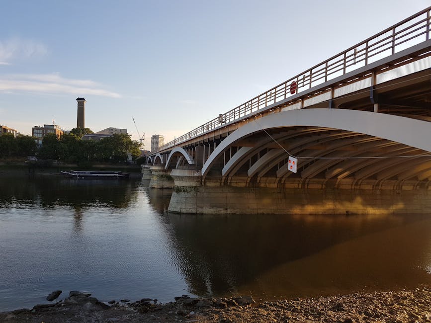 A wide view of Kingston Bridge spanning the River Thames in Kingston upon Thames during daytime, with the bridge's white arch supports and metal railings visible along its length. The river below reflects the structure and nearby trees, which line the riverbank on the far side. In the background, a tall chimney and various mid-rise buildings, including a construction crane, are visible under a partly cloudy sky with sunlight illuminating the scene. The foreground shows the rocky shoreline with some small stones, indicating the edge of the riverbank. This scene is related to home relocation and moving logistics, illustrating the vicinity around Kingston Bridge where vehicle access is necessary for property removals, such as loading and unloading furniture and packing materials. For professional removals in Kingston upon Thames, services often consider local access points like this, guided by infrastructure such as the bridge, to facilitate efficient furniture transport during house removals conducted by companies like manwithvankingstonuponthames.org.uk.