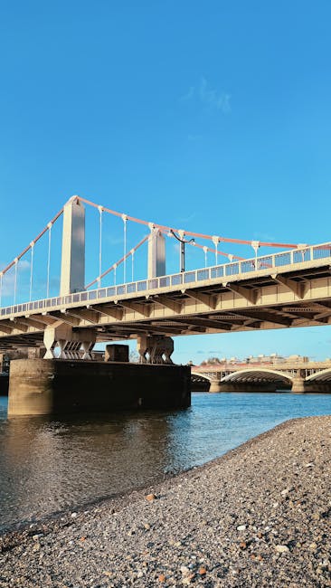 A clear view of Kingston Bridge spanning the River Thames under a bright blue sky, with the suspension bridge's white cables and metal structure prominently visible. Below, calm river waters reflect the bridge and the surrounding environment. The shoreline in the foreground consists of small pebbles and gravel, indicating a riverside location. In the distance, additional bridge structures are visible along the river. This scene is typical of the area around Kingston upon Thames, a popular location for house removals and furniture transport services, as evidenced by the bridge's access points suitable for vans and removal trucks. The image captures the outdoor environment where loading and unloading of furniture and household items may occur during home relocation or moving logistics, consistent with professional removals operations carried out by companies such as those detailed in the Kingston Bridge removals access guide for vans.