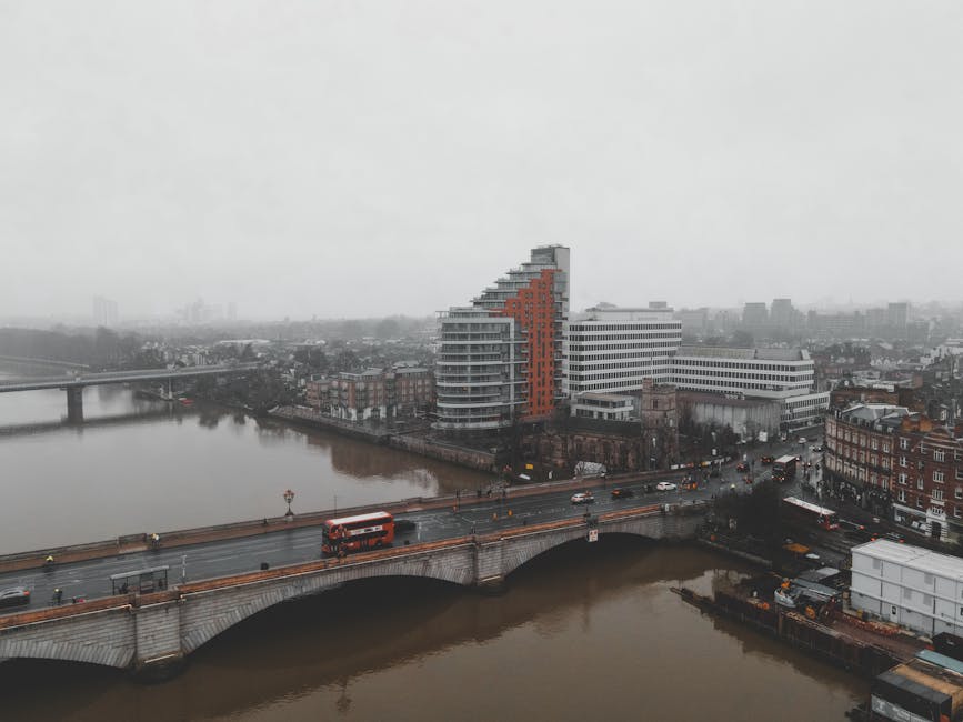 A wide view of Kingston Bridge spanning the River Thames in Kingston upon Thames during daytime, with the bridge's white arch supports and metal railings visible along its length. The river below reflects the structure and nearby trees, which line the riverbank on the far side. In the background, a tall chimney and various mid-rise buildings, including a construction crane, are visible under a partly cloudy sky with sunlight illuminating the scene. The foreground shows the rocky shoreline with some small stones, indicating the edge of the riverbank. This scene is related to home relocation and moving logistics, illustrating the vicinity around Kingston Bridge where vehicle access is necessary for property removals, such as loading and unloading furniture and packing materials. For professional removals in Kingston upon Thames, services often consider local access points like this, guided by infrastructure such as the bridge, to facilitate efficient furniture transport during house removals conducted by companies like manwithvankingstonuponthames.org.uk.