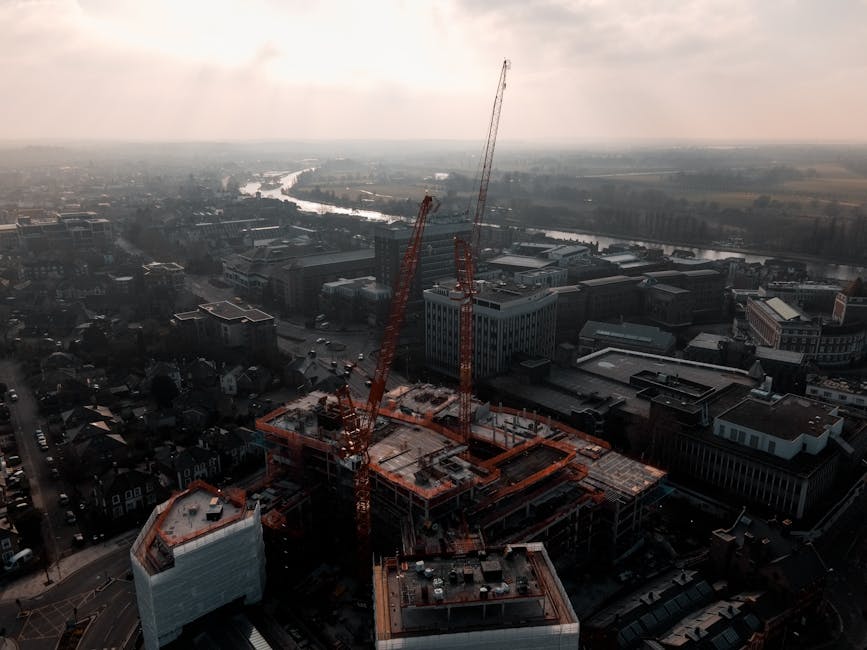 Aerial view of a construction site with three tall orange cranes positioned around a partially built multi-storey building, situated in an urban area with surrounding office buildings and residential