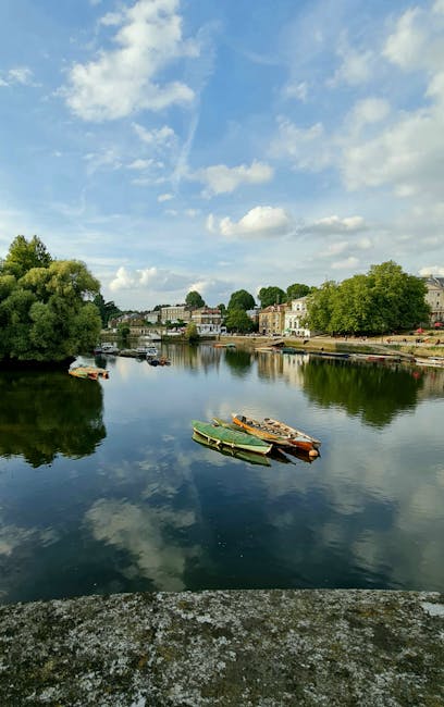 A calm river scene during daytime with a partly cloudy sky overhead. Several small boats, including green and brown ones, are moored along the riverbank. On the opposite side of the river, there are residential buildings and trees, some with lush green foliage. The water reflects the sky, clouds, and surrounding greenery, creating a tranquil atmosphere. This setting is typical of Kingston upon Thames, where house removals and furniture transport services such as those provided by manwithvankingstonuponthames.org.uk may involve navigating scenic waterways during home relocation or moving processes.