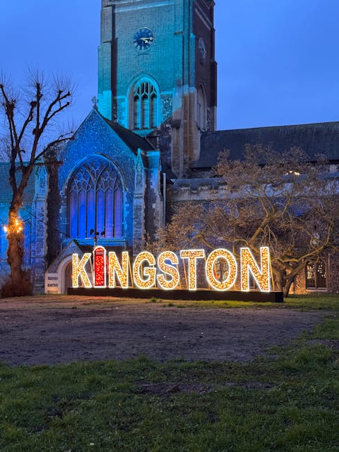 A large, illuminated sign spelling 'KINGSTON' in bright red and white lights is situated on a grassy area in front of a historic stone church with tall, arched windows. The church tower, constructed from grey stone, rises prominently behind the sign and features a clock face and pointed Gothic-style windows, illuminated with blue lighting at dusk. To the right of the sign, there is a leafless tree with twisted branches, and to the left, a streetlamp casts a warm glow. The scene captures a moment during an active home relocation process, with a partially visible van and moving blankets on the pavement nearby, indicating furniture transport and packing activities typical of house removals in Kingston upon Thames. This setting reflects the local environment where [COMPANY_NAME] provides professional removals and moving services, supporting smooth home relocation logistics.
