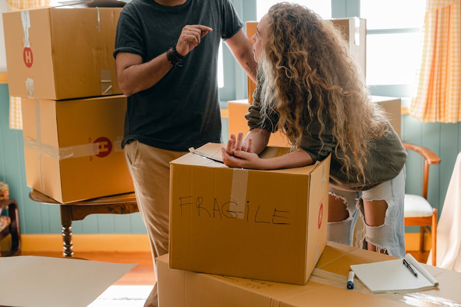 A woman with long, curly blonde hair leaning on a cardboard box labeled 'FRAGILE' during a home relocation, with her arms resting on the box and her head turned to the side. She is inside a room with green-paneled walls and yellow checkered curtains. To her right, a man wearing a black T-shirt and beige pants is partially visible, appearing to be assisting with packing or loading. Several other cardboard boxes, some stacked on a wooden table in the background, are sealed with packing tape and marked with symbols. The scene is well-lit with natural light coming through the windows, emphasizing the ongoing packing process as part of furniture transportation and moving logistics handled by [COMPANY_NAME]. A clipboard and a pen are placed on the table nearby, indicating organisation and planning during the packing and loading process for house removal services.