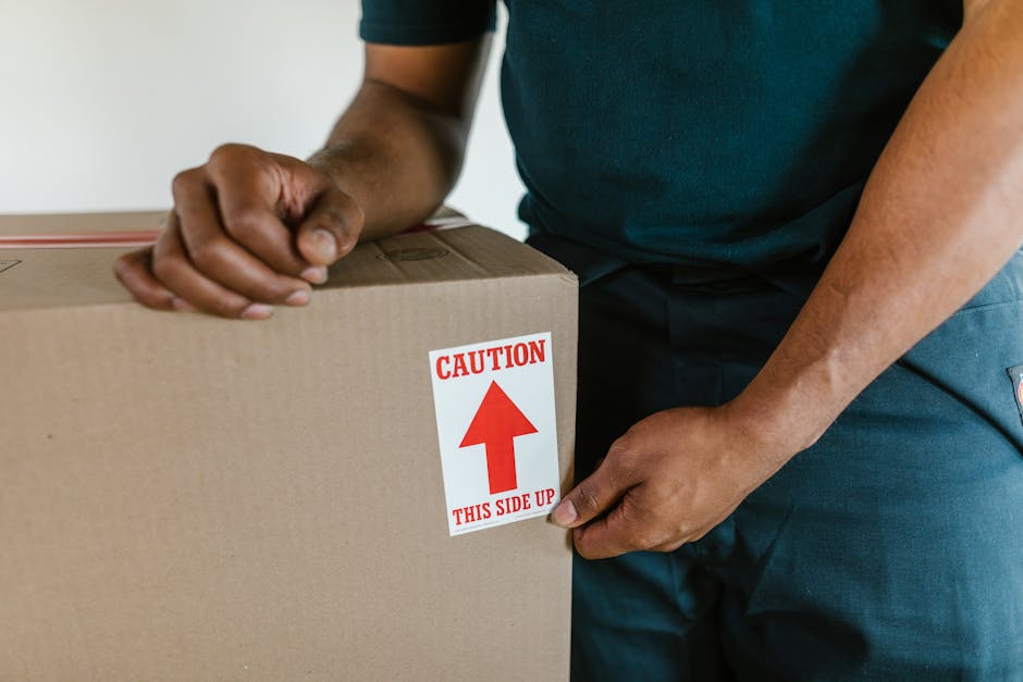 A person wearing a dark green t-shirt and blue work trousers is seen holding a large cardboard box indoors, with one hand gripping the top edge and the other supporting the bottom. The box features a white label with red text and a red upward-pointing arrow, warning 'CAUTION THIS SIDE UP'. The setting appears to be a home environment, potentially a hallway or living room, with a neutral background. This image illustrates careful handling during packing in a home relocation, highlighting important packaging instructions used by the Kingston home removals teams, such as proper orientation to prevent damage during furniture transport and loading processes.