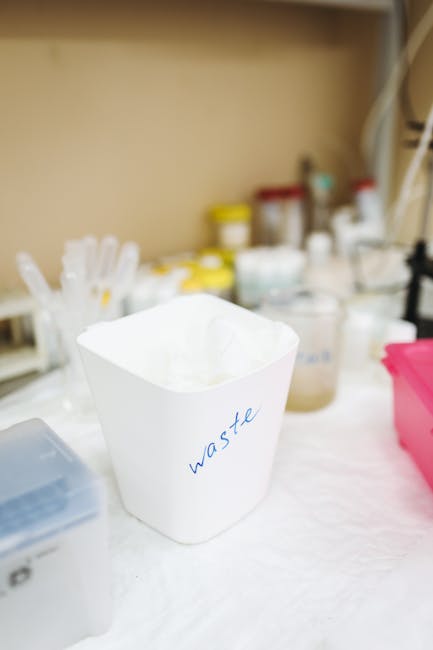 A close-up view of a white paper waste bin labeled 'waste' in blue ink, placed on a white surface inside a room used for packing and packing materials. The background features various containers, bott